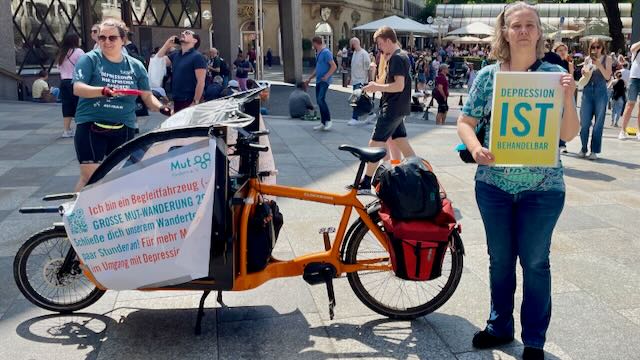 Mut-Tour-Wanderung 2025: Zwei Teilnehmerinnen mit Lastenrad und Schildern "Depression ist behandelbar" und Ich bin ein Begleitfahrzeug der GROSSEN MUT-WANDERUNG" auf der Kölner Domplatte