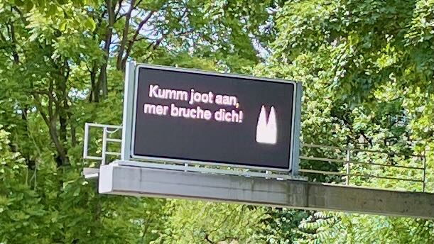 Straßenschild in Köln mit der Aufschrift "Kumm joot aan, mer bruche dich" (auf Hochdeutsch: "Komm gut an, wir brauchen dich!"